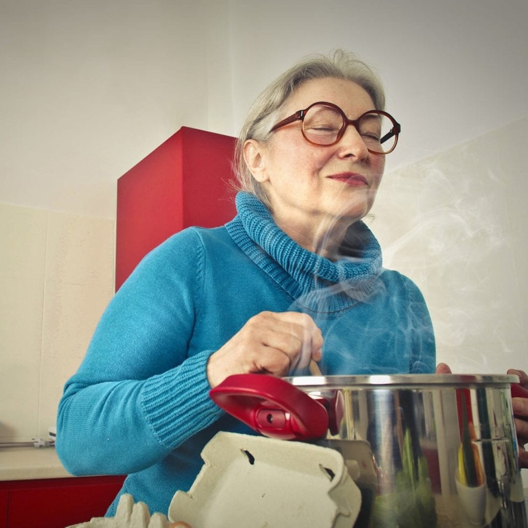 A elderly lady wearing glasses cooks over a pot of soup, smiling and enjoying the smell.