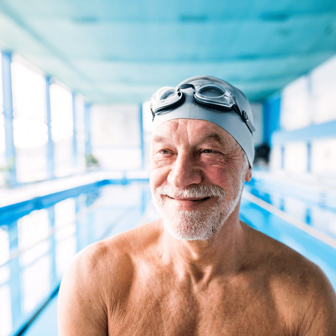 A senior wearing a swim cap and goggles smiles with a pool in the background.